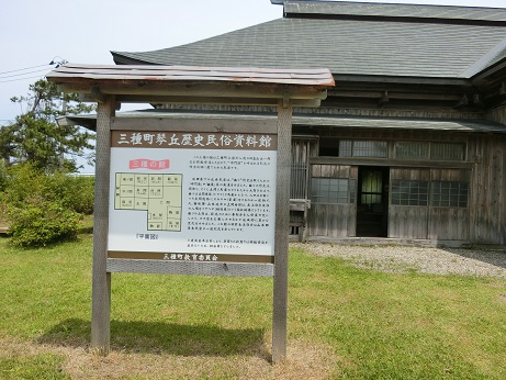 三種町琴丘歴史民俗資料館の案内版の後ろに、屋根が銅板葺となっており、木造の古い平屋の建物、三種の館の外観写真