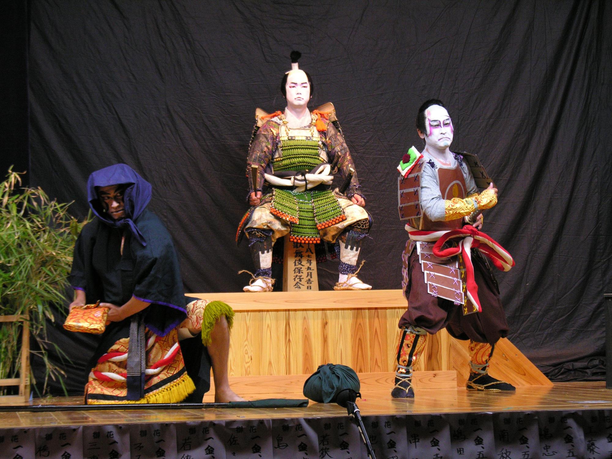 歌舞伎の衣装を着て、隈取をした3人の人物がおり、2人は立ってポーズをとり、もう1人は中央の一段高いところで椅子に腰かけ座っている歌舞伎のシーンの写真
