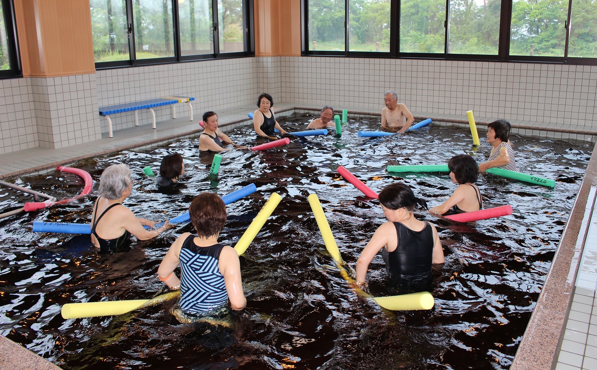 参加者達がプールの中で大きな円になり、アクアポールをそれぞれ持って温泉浴運動を行っている写真