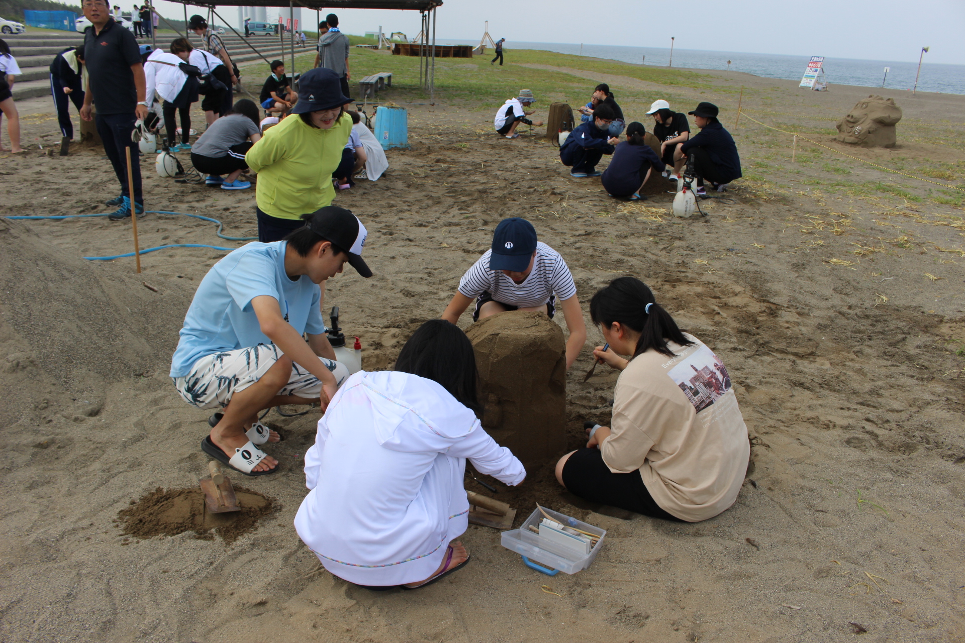 海水浴場の浜辺に座り砂像を作る男女のグループと、指導する講師の写真