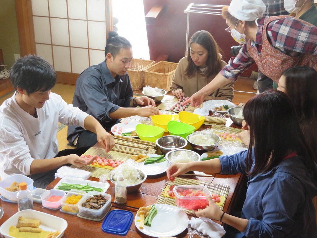 テーブルの上に材料を並べて、飾り巻きずし作りをしている若い男女のグループと、指導をする年配の女性の写真