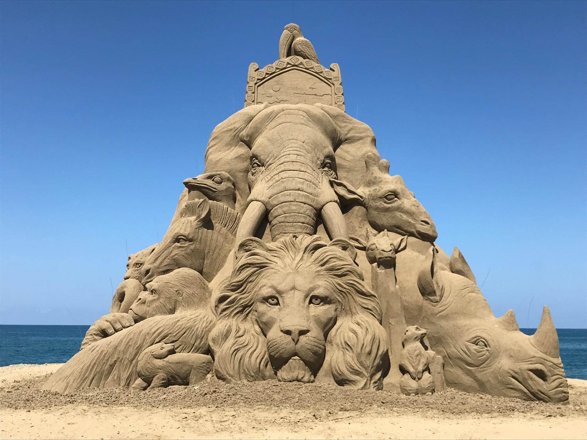 正面に像とライオンの顔があり、サイやゴリラ、馬など沢山の動物たちが彫刻された海辺の砂浜に作られた巨大で複雑な砂の彫刻の写真